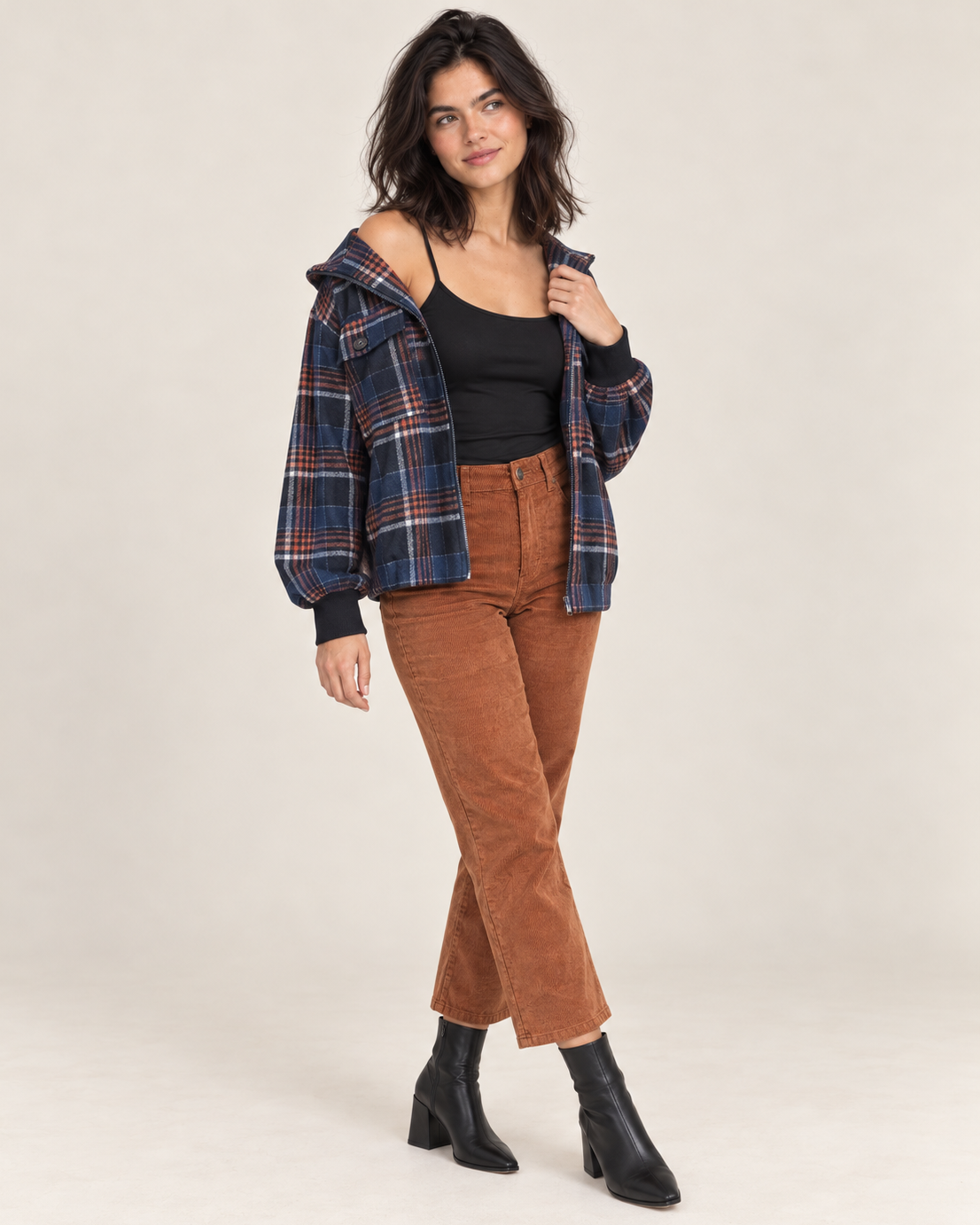 Full-body front view of a woman wearing a black spaghetti-strap camisole with a navy and rust plaid jacket, rust brown flared pants, and black heeled ankle boots on a beige background