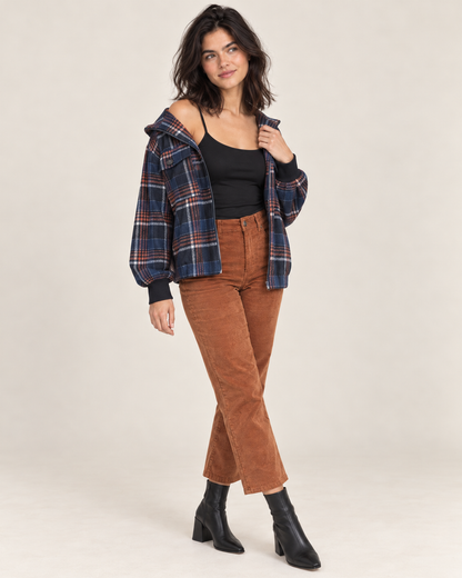 Full-body front view of a woman wearing a black spaghetti-strap camisole with a navy and rust plaid jacket, rust brown flared pants, and black heeled ankle boots on a beige background