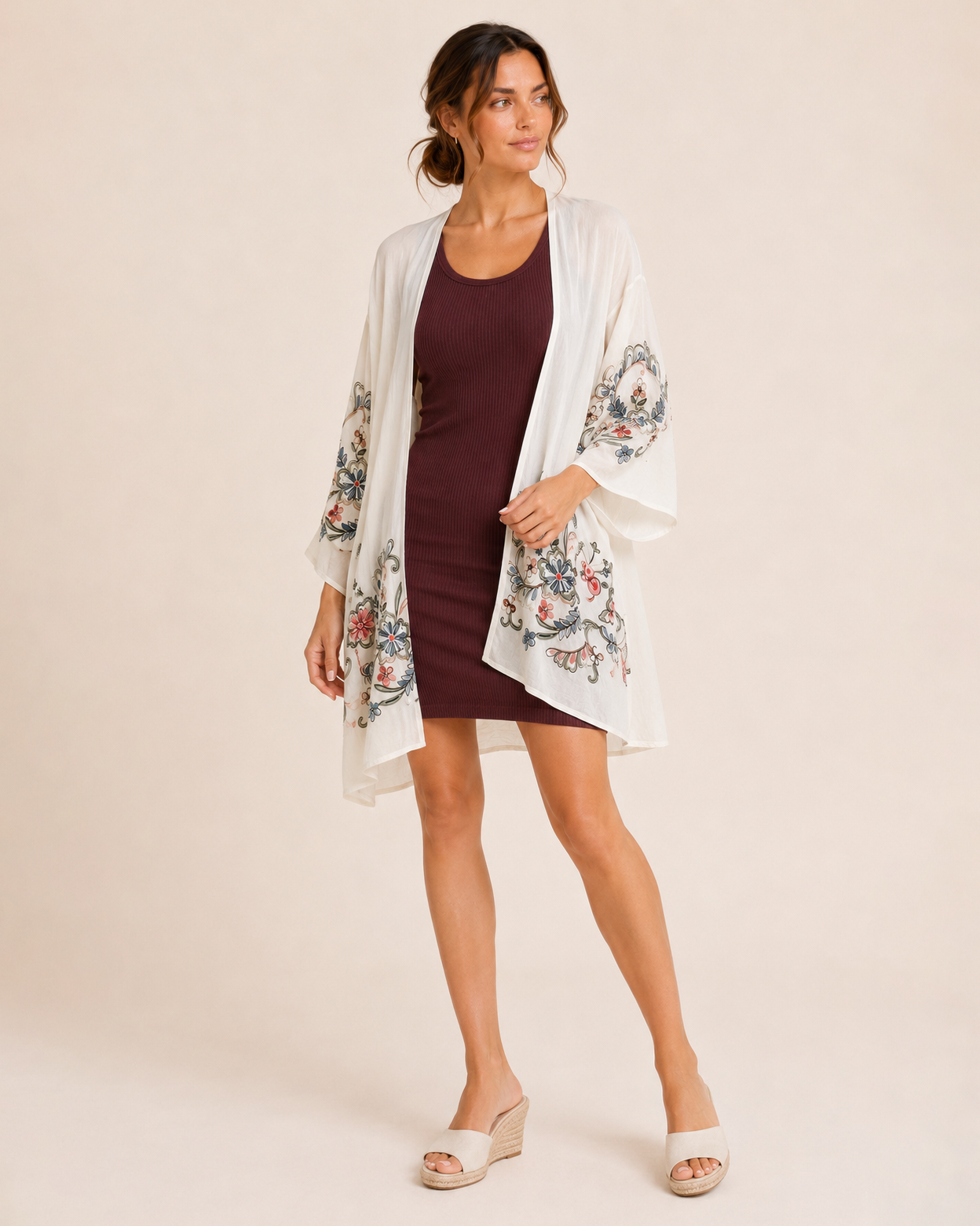 Full-body view of a woman wearing a cream floral kimono over a burgundy ribbed tank dress and brown sandals on a beige background
