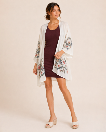 Full-body view of a woman wearing a cream floral kimono over a burgundy ribbed tank dress and brown sandals on a beige background
