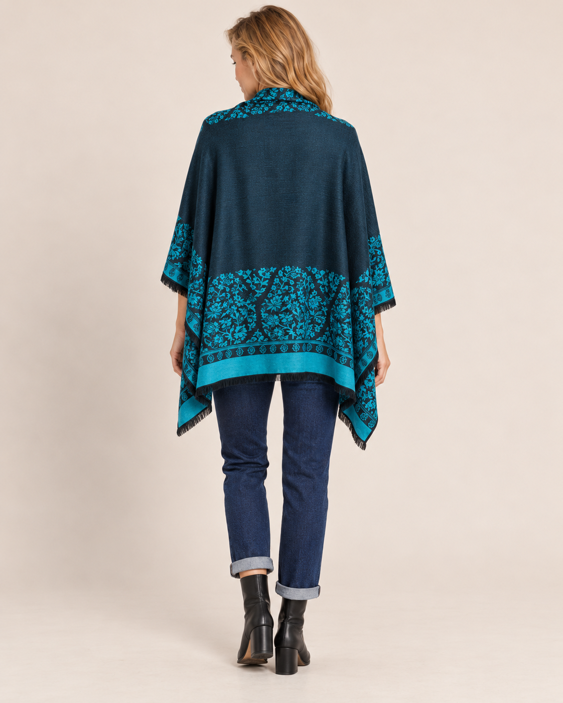Back view of a woman wearing a teal and black Southwestern geometric-patterned poncho cardigan over a dark top, paired with dark blue skinny jeans and black ankle boots on a beige background.