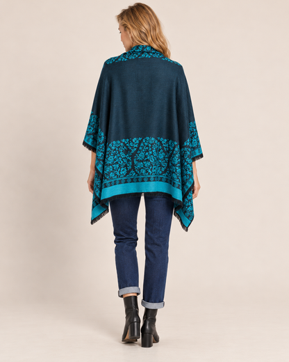 Back view of a woman wearing a teal and black Southwestern geometric-patterned poncho cardigan over a dark top, paired with dark blue skinny jeans and black ankle boots on a beige background.