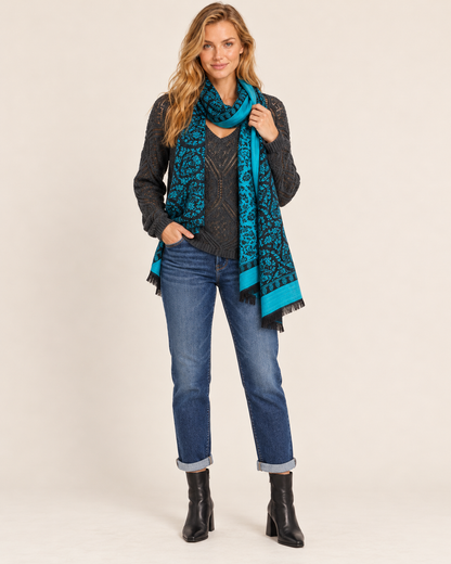 Three-quarter front view of a woman wearing a teal and black Southwestern geometric-patterned poncho cardigan over a dark top, paired with dark blue skinny jeans and black ankle boots on a beige background.