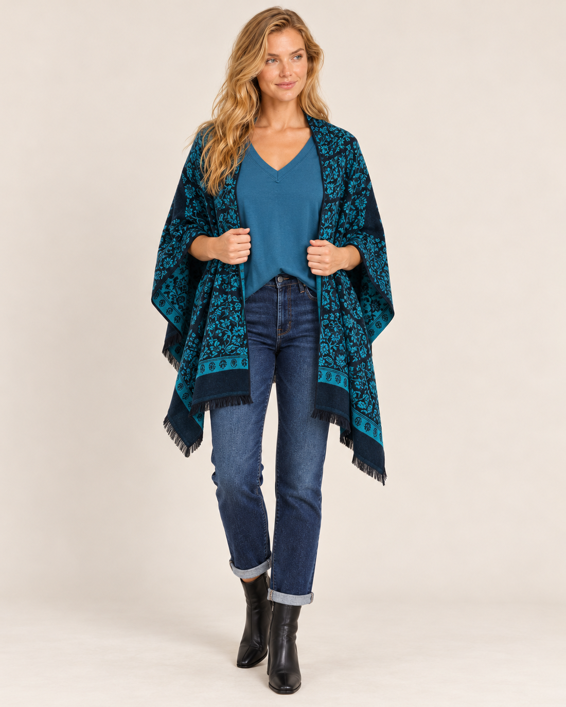 Full-body front view of a woman wearing a teal and black Southwestern geometric-patterned poncho cardigan over a dark top, paired with dark blue skinny jeans and black ankle boots on a beige background.