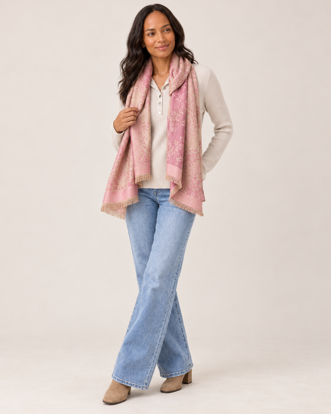 Full-body side view of a woman wearing a pink patterned open-front ruana wrap over a white V-neck top, paired with light-wash flared jeans and white sneakers on a beige background.