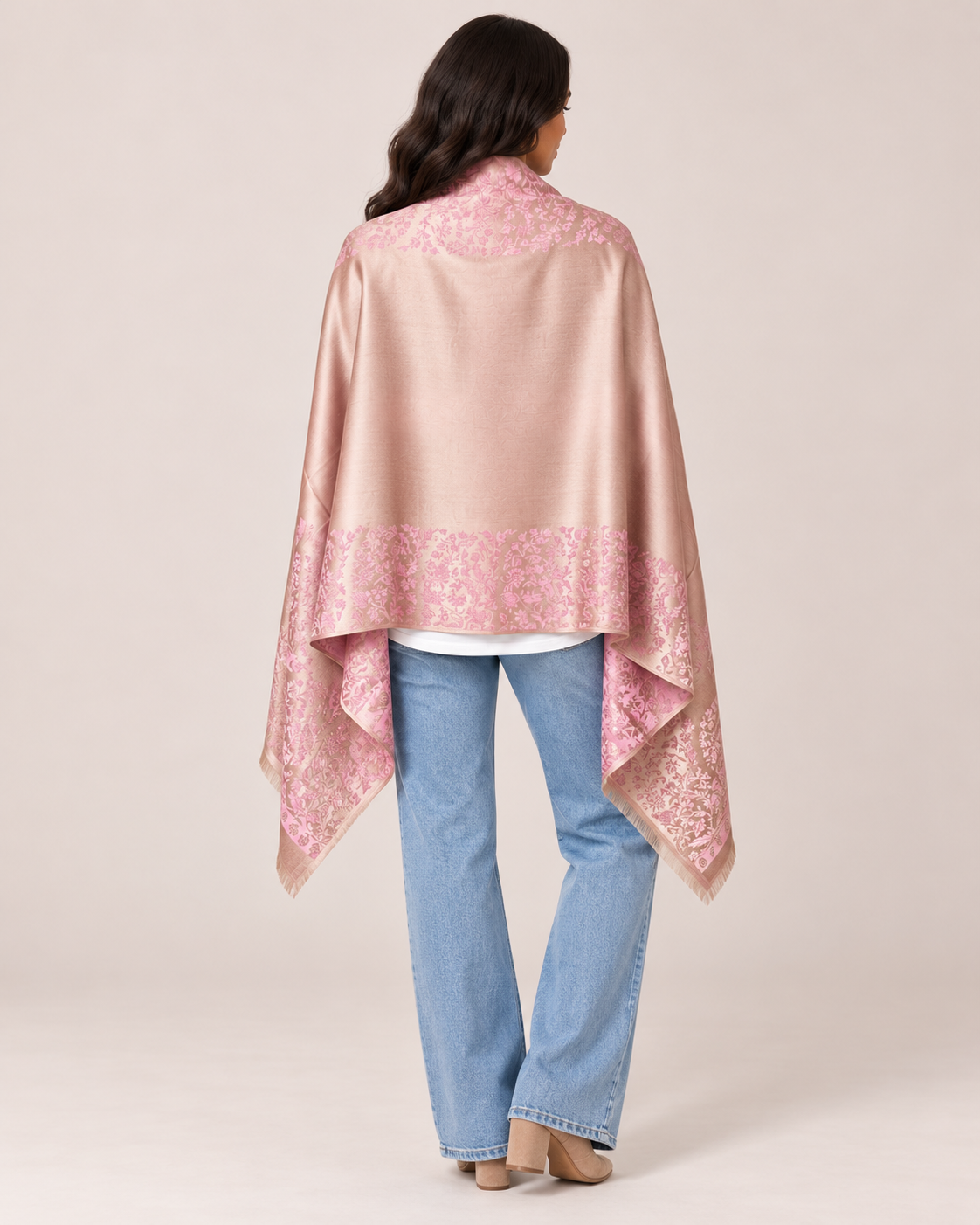 Back view of a woman wearing a pink patterned ruana wrap with a draped silhouette over light-wash jeans on a beige background.