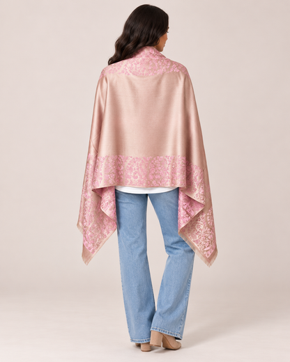 Back view of a woman wearing a pink patterned ruana wrap with a draped silhouette over light-wash jeans on a beige background.