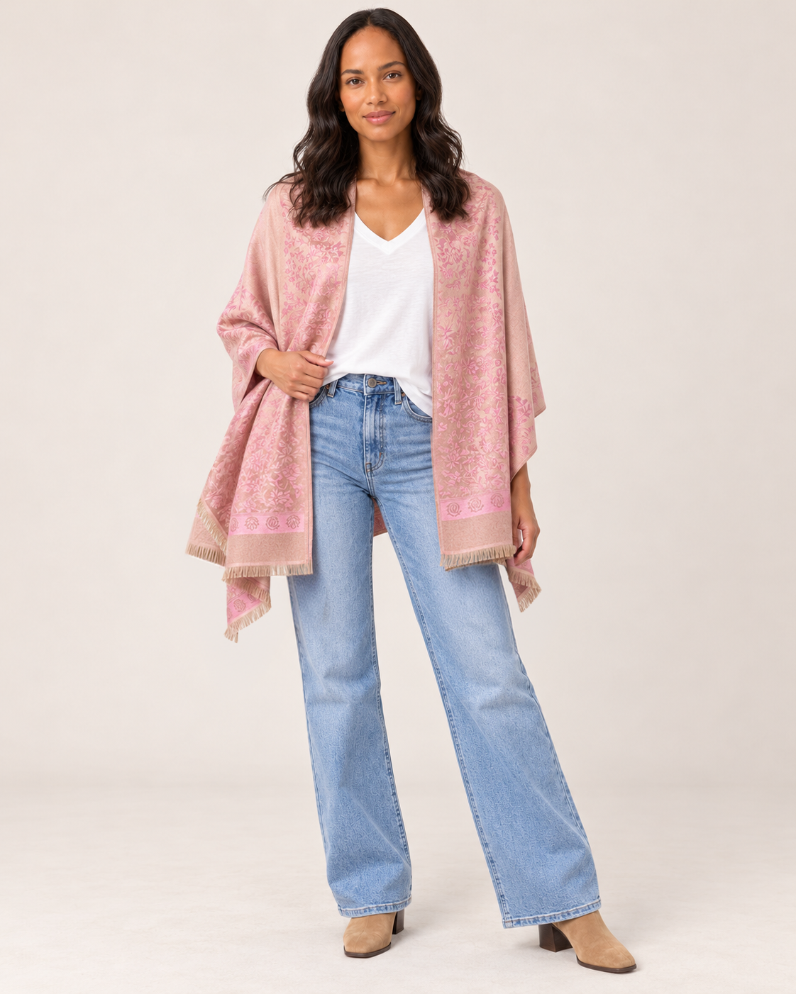 Full-body front view of a woman wearing a pink patterned open-front ruana wrap over a white V-neck top, paired with light-wash flared jeans and white sneakers on a beige background.