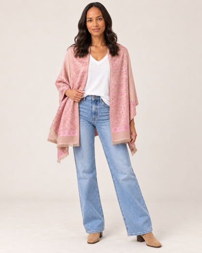 Full-body front view of a woman wearing a pink patterned open-front ruana wrap over a white V-neck top, paired with light-wash flared jeans and white sneakers on a beige background.