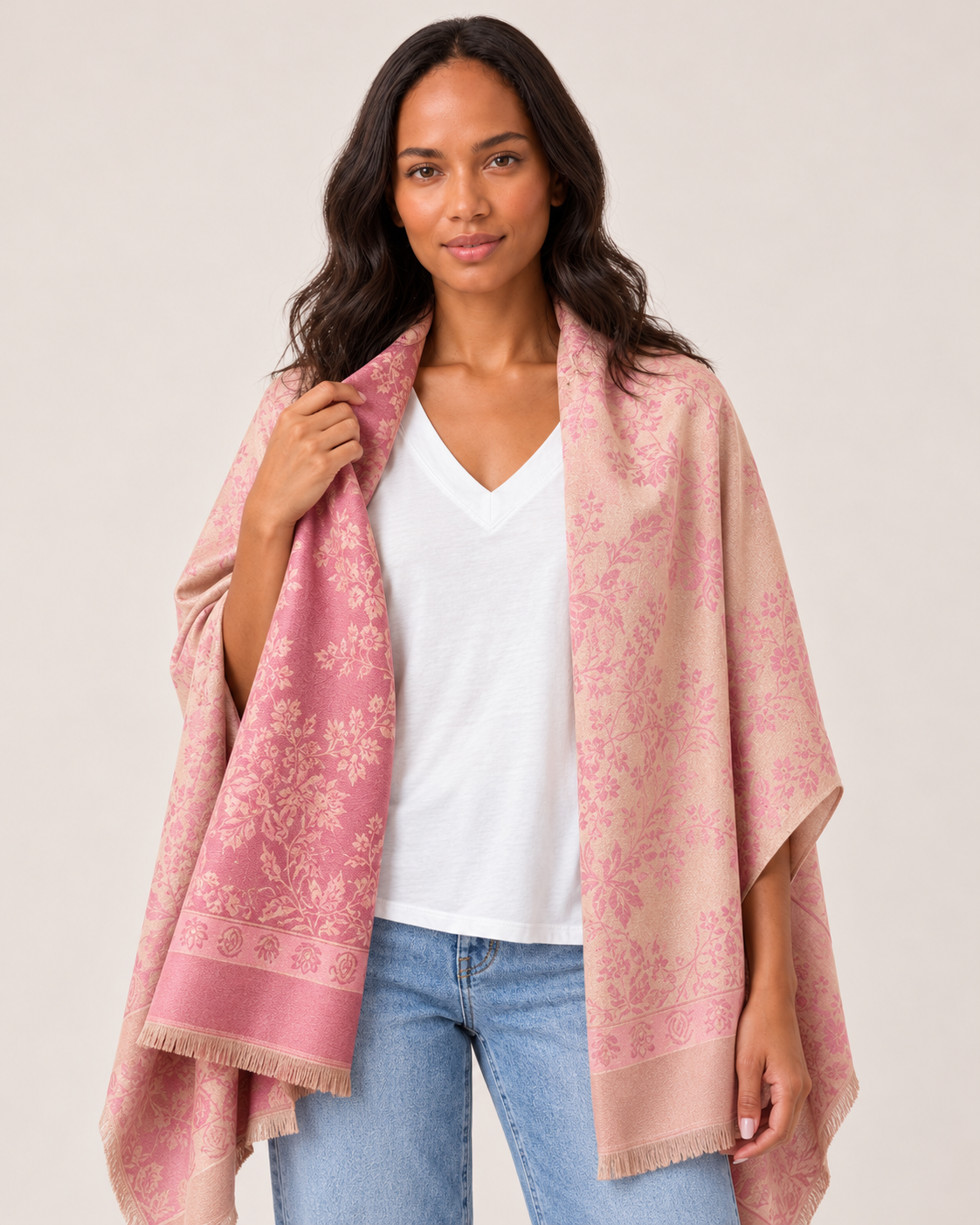 Cropped front view of a woman wearing a pink patterned ruana wrap over a white V-neck top and light-wash jeans, highlighting the draped design and woven pattern on a beige background.
