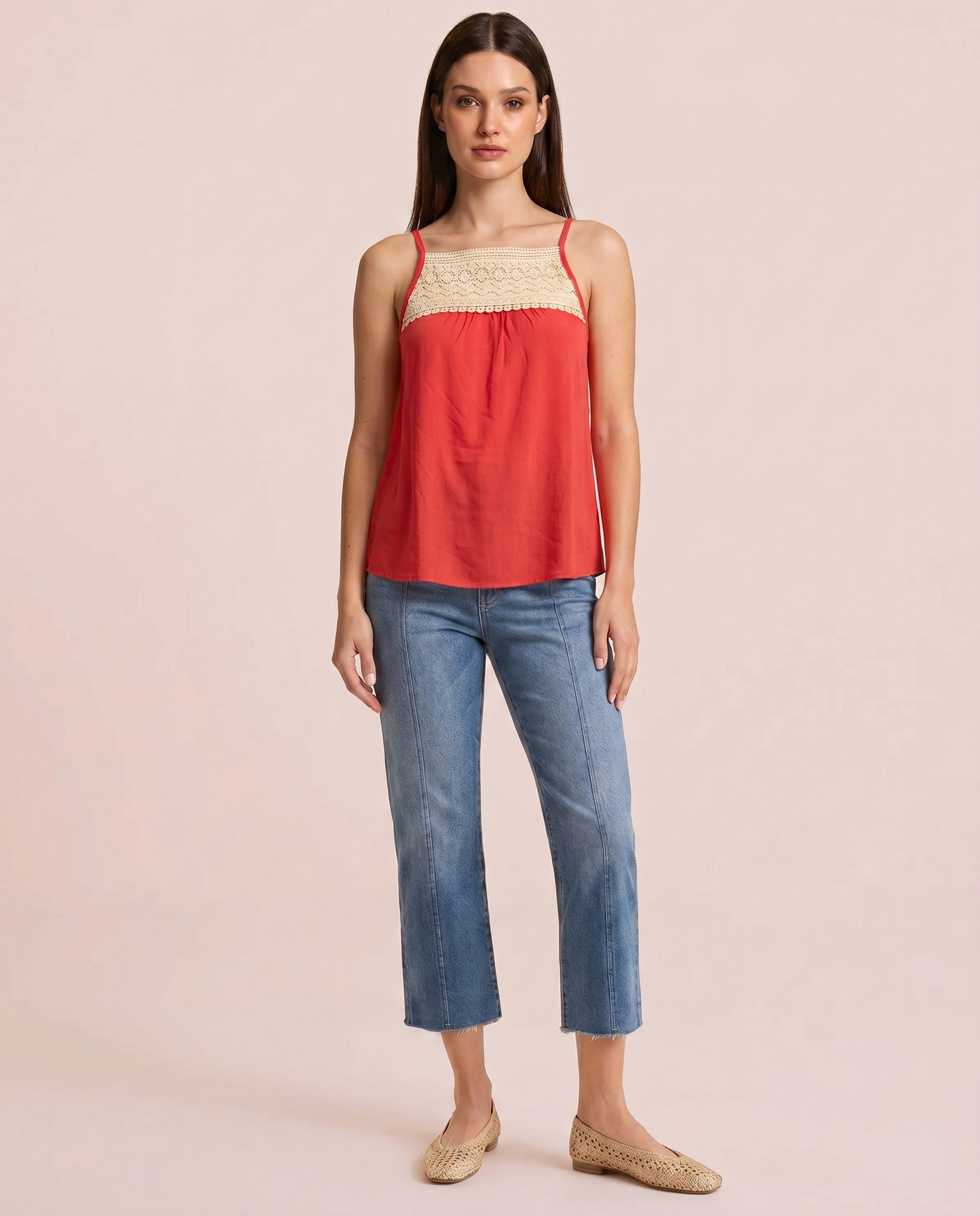 Front view of a woman wearing a coral camisole top with a crochet lace yoke and blue jeans on a beige background