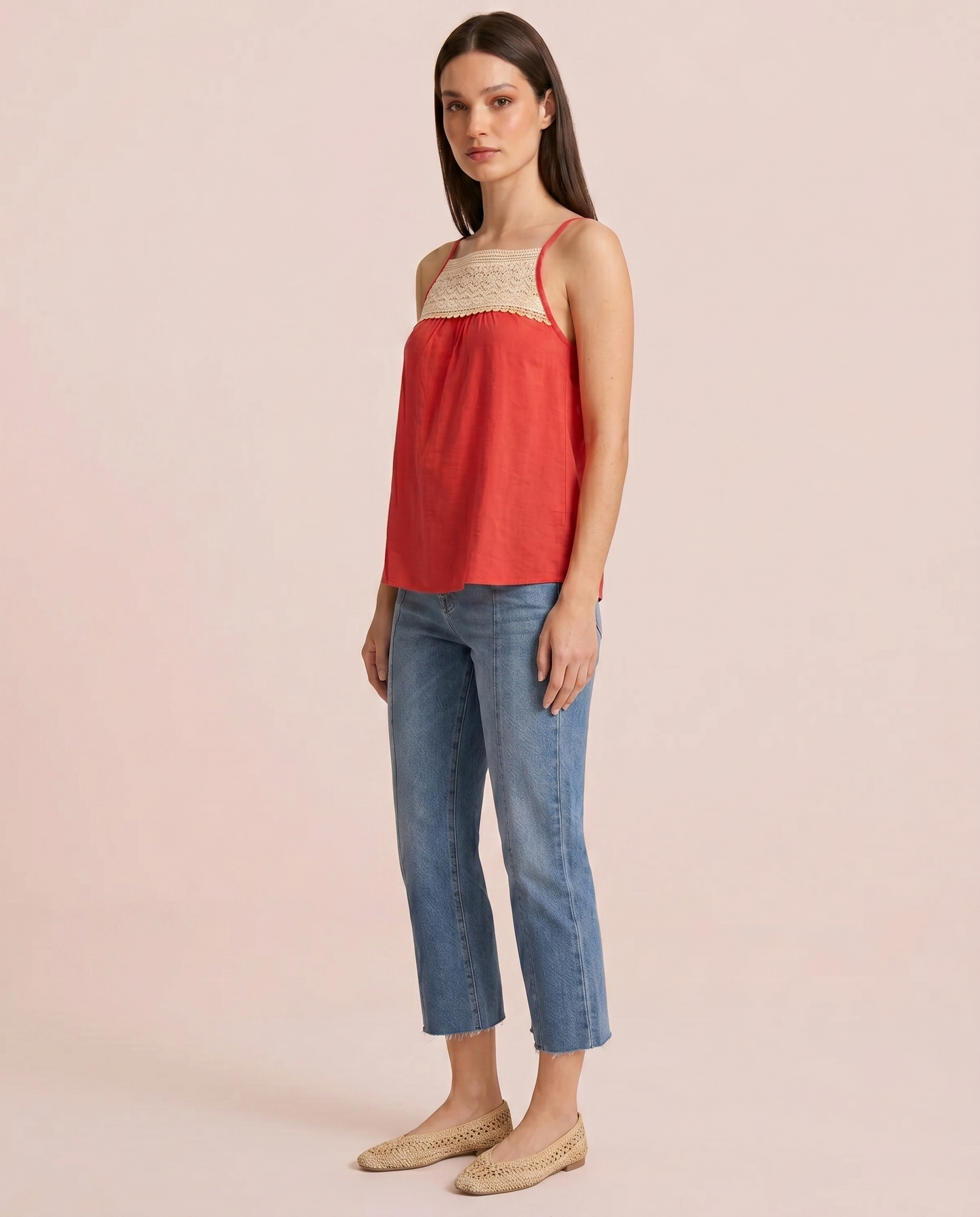 Side view of a woman wearing a coral camisole top with a crochet lace yoke and blue jeans on a beige background