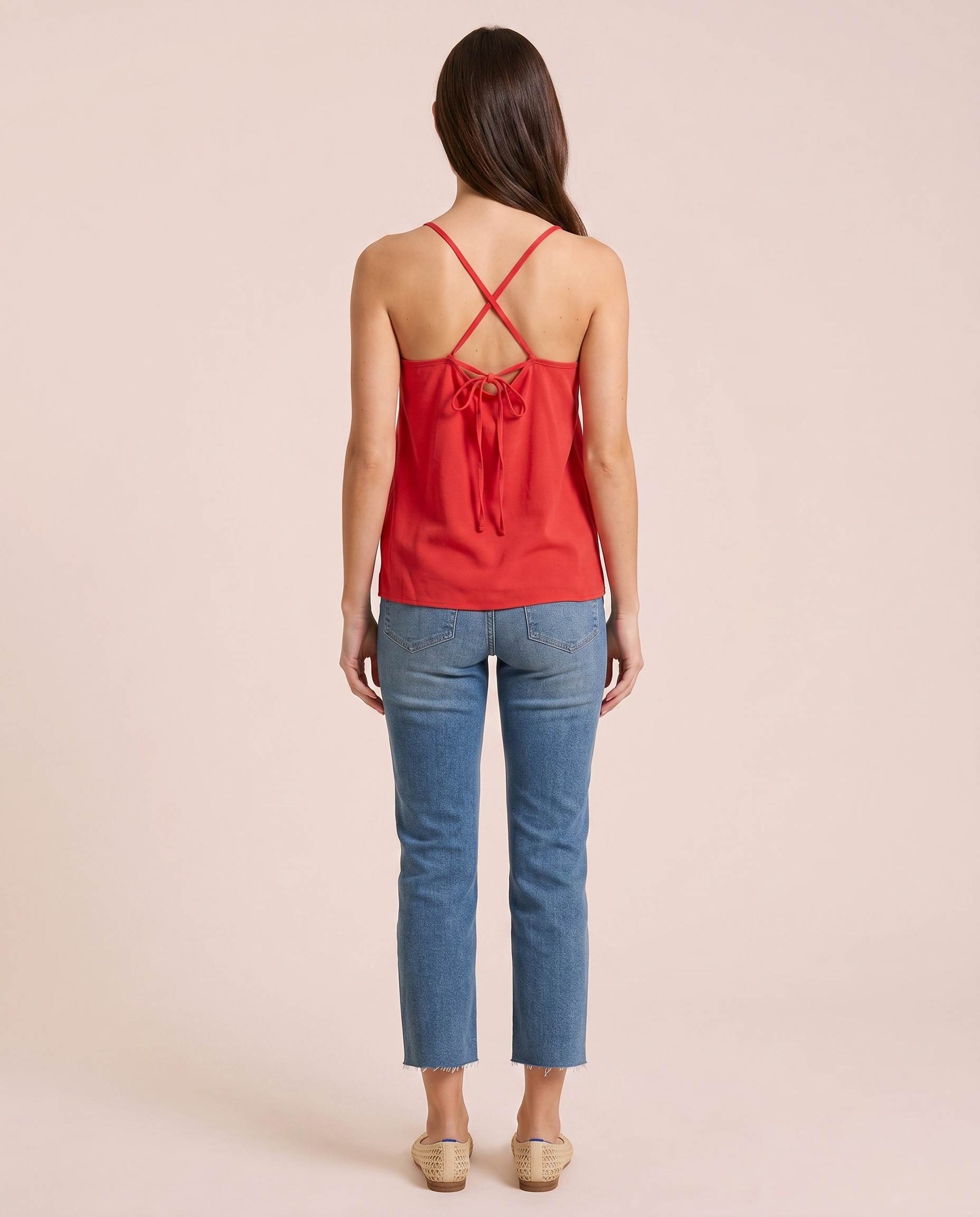 Back view of a woman wearing a coral camisole top with crisscross spaghetti straps and blue jeans on a beige background
