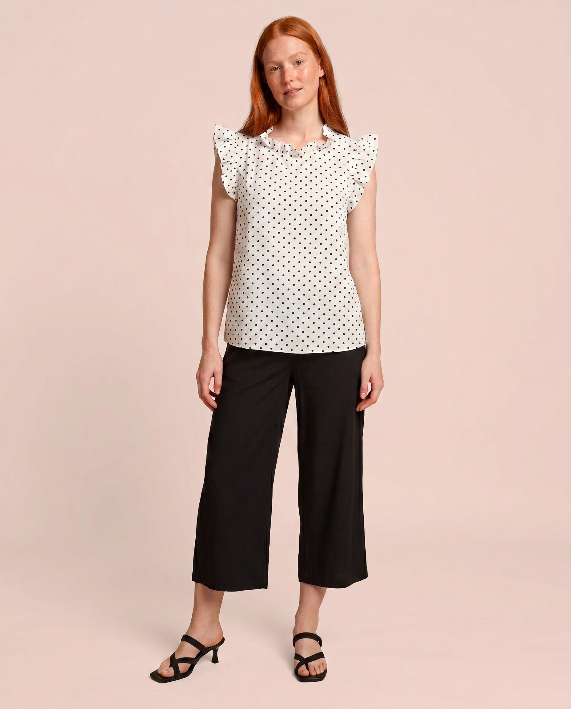 Front view of a woman wearing a white blouse with black polka dots and black pants on a beige background