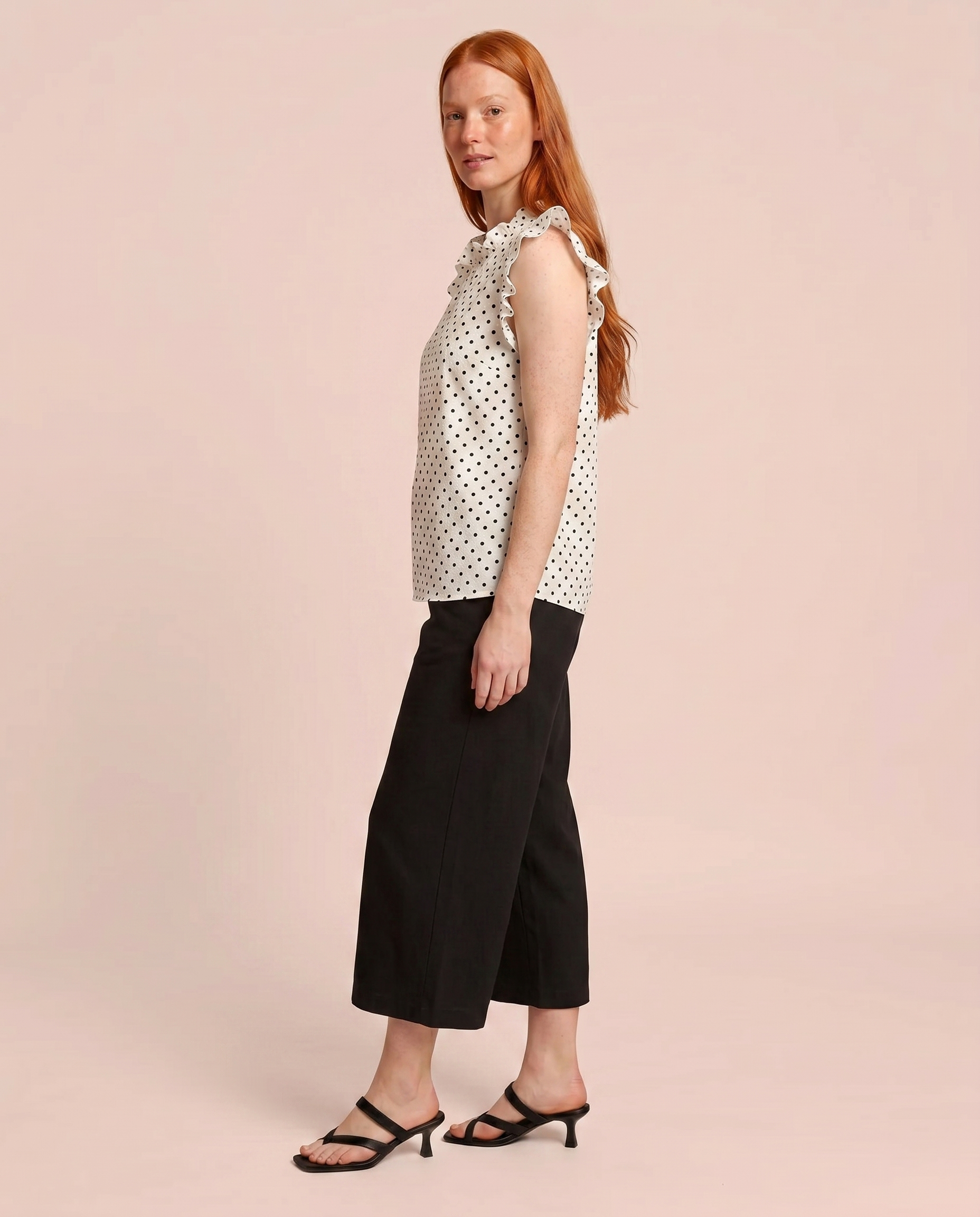 Side view of a woman wearing a white blouse with black polka dots and black pants on a beige background