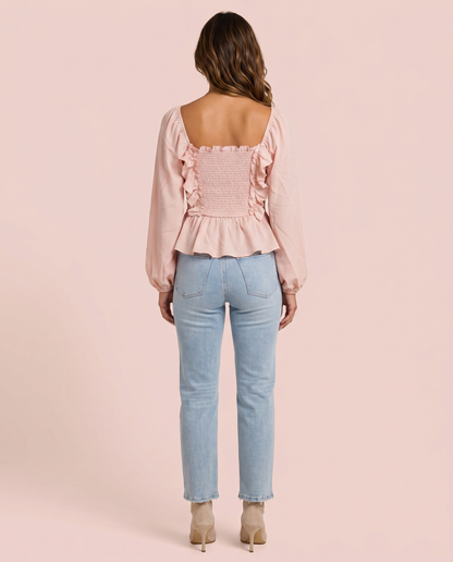 Back view of woman wearing a pink ruffled peplum blouse and light blue jeans on a pink background