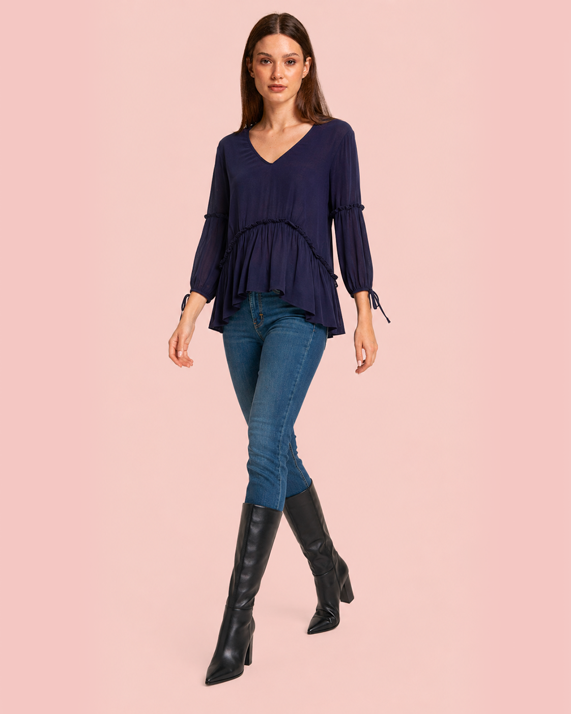 Woman wearing a navy blue top, jeans, and black knee-high boots on a pink background