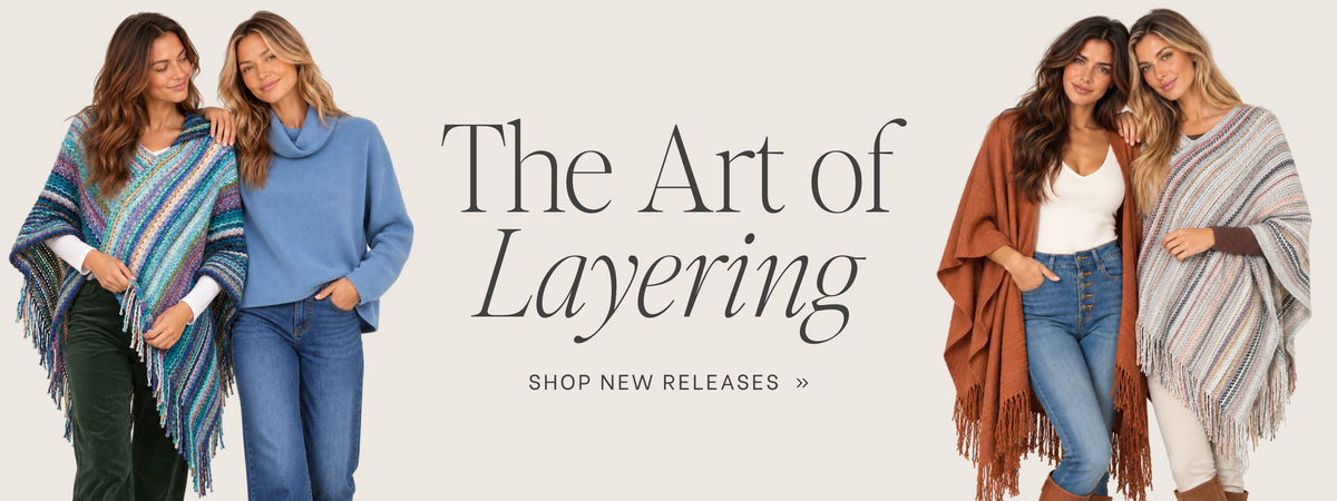 A promotional banner with the text 'The Art of Layering' and 'SHOP NEW RELEASES' on a light beige background. Three women are shown wearing layered clothing and shawls: a blue sweater with jeans on the left, a rust-brown fringed shawl over a white top and jeans in the center, and a gray-and-white striped shawl over a white top and white pants on the right.
