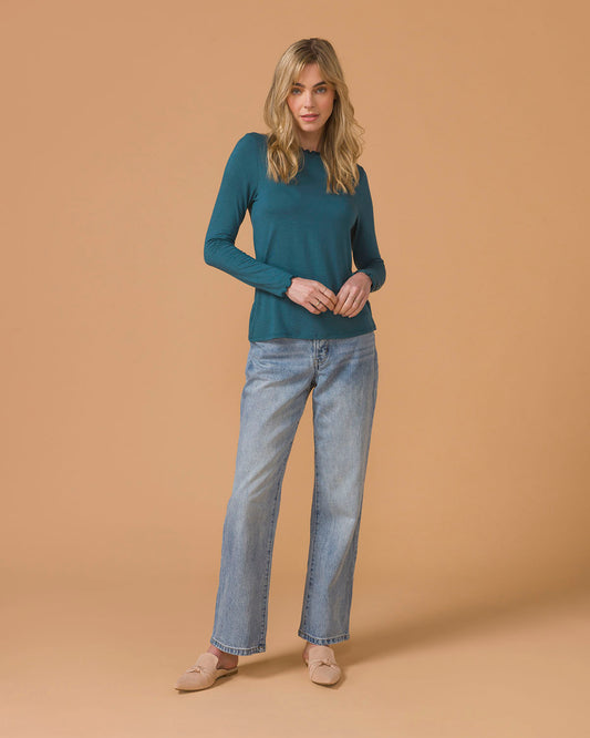 Woman wearing a teal long-sleeve top and light blue jeans sitting on a stool