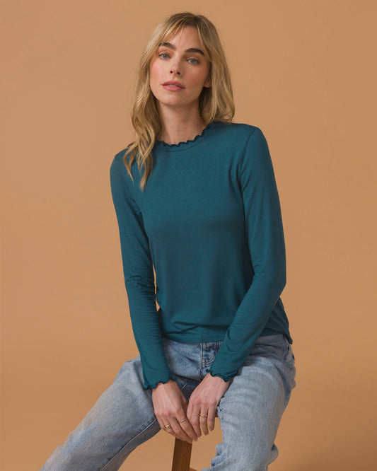 Woman wearing a teal long-sleeve top and light blue jeans sitting on a stool