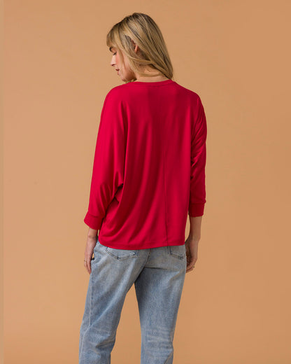 Woman wearing a red long sleeve top and blue jeans against a beige background back view