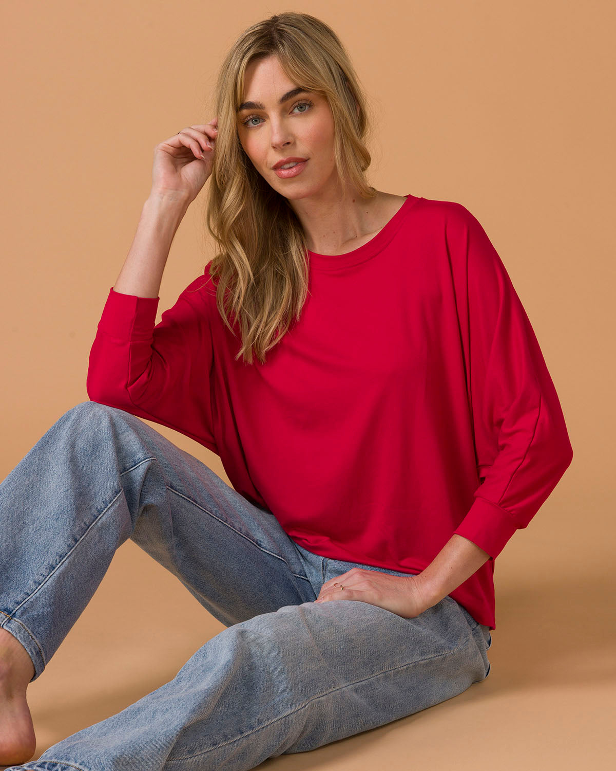 Seated woman wearing a red long sleeve top and blue jeans on a beige background