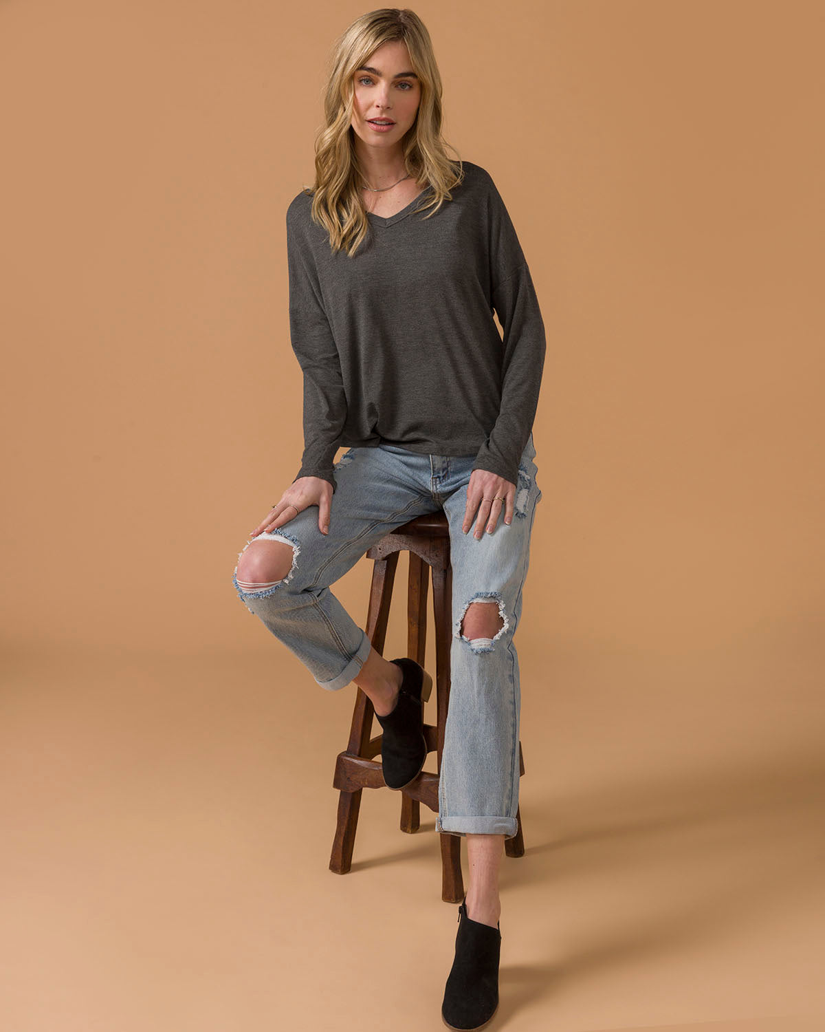 Woman sitting on a stool wearing a dark gray long sleeve top and light blue jeans with a brown background