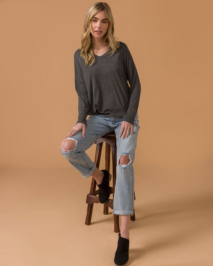 Woman sitting on a stool wearing a dark gray long sleeve top and light blue jeans with a brown background