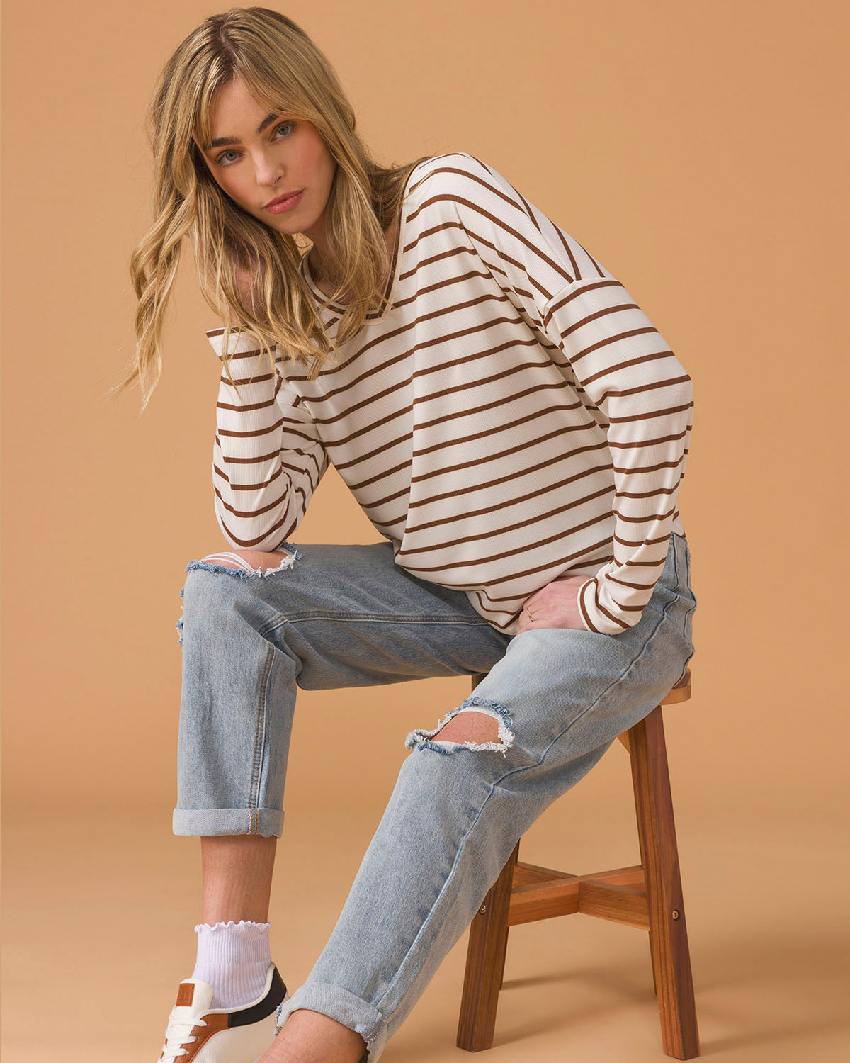 Woman wearing a striped long sleeve top and blue jeans sitting on a stool on a beige background