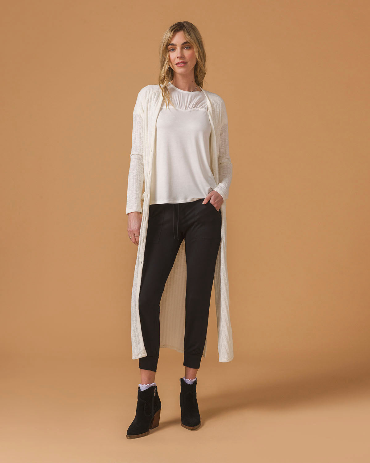 Woman wearing a long cream cardigan, white top, black pants on a beige background