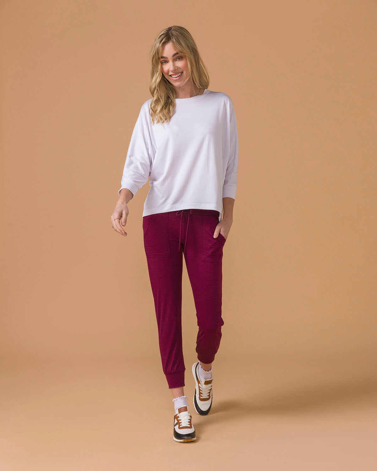 Woman wearing a white long sleeve top and maroon  jogger pants on a beige background