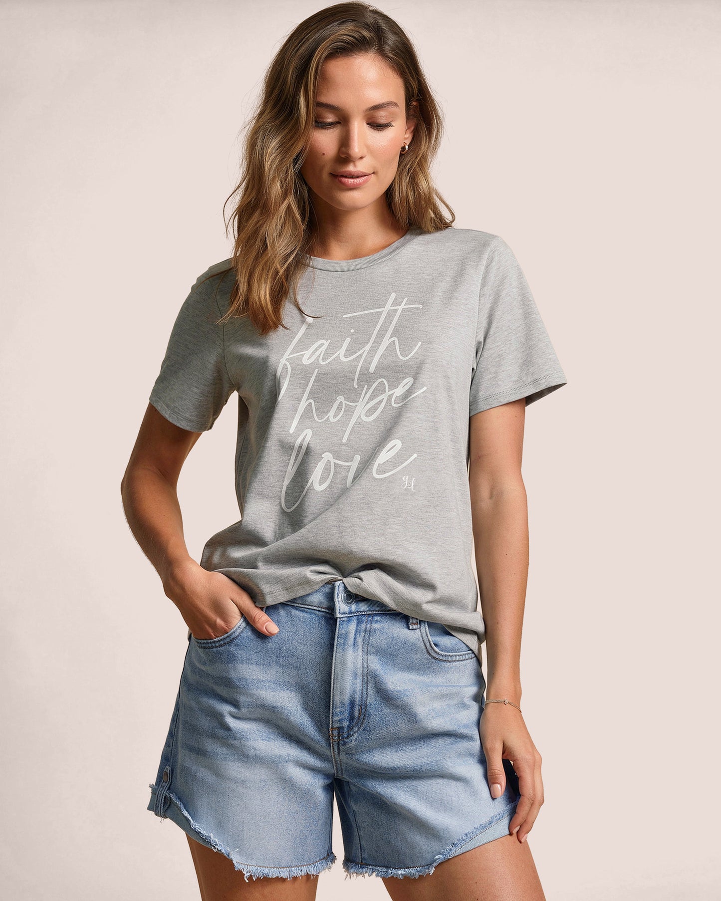 Woman wearing a gray t-shirt with 'faith hope love' text and denim shorts on a plain background