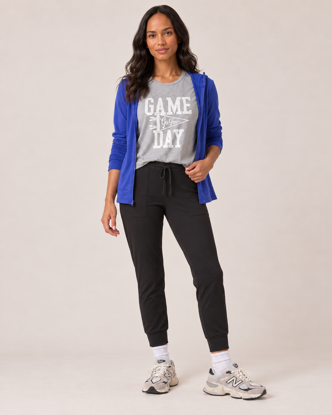 A woman modeling a gray graphic t-shirt with 'GAME 4 DAY' in white, layered under a royal blue zip-up hoodie, paired with black jogger pants, white socks, and New Balance sneakers against a light beige studio background.