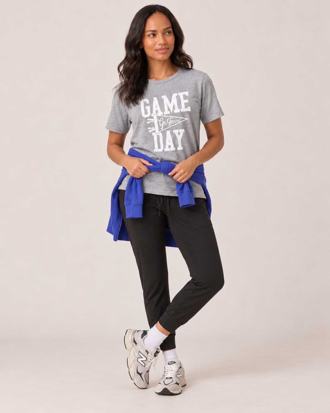 A woman modeling a heather gray graphic T-shirt with 'GAME DAY' printed in white, wearing black joggers, white socks, and New Balance sneakers, while holding a blue shirt tied around her waist against a plain light beige studio background.
