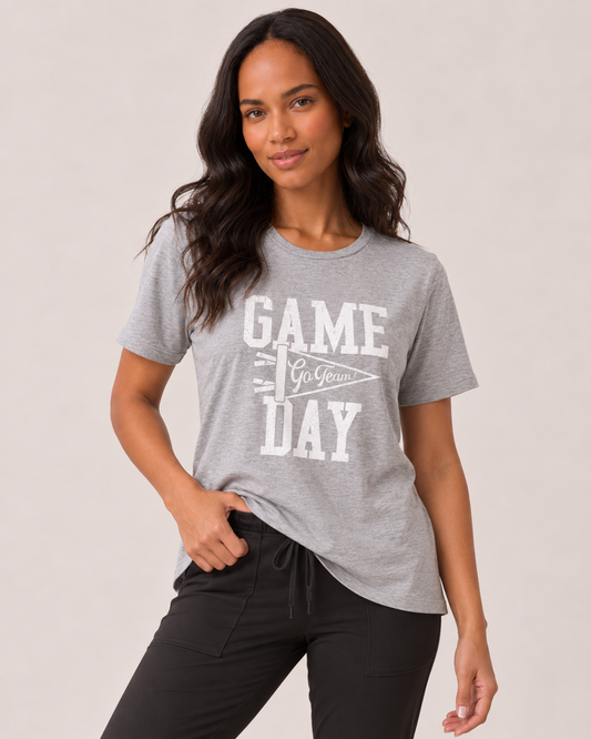 A woman modeling a heather gray short-sleeve t-shirt with a white graphic that reads 'GAME DAY' and 'Go Yeezus'. She is wearing black drawstring pants against a plain light beige studio background.