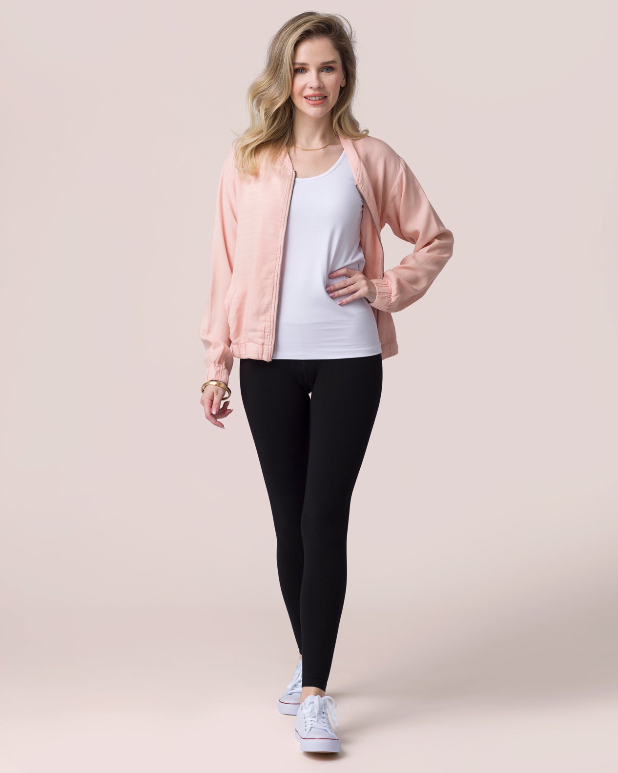 Woman wearing a pink jacket, white top, black leggings, and blue sneakers on a light pink background
