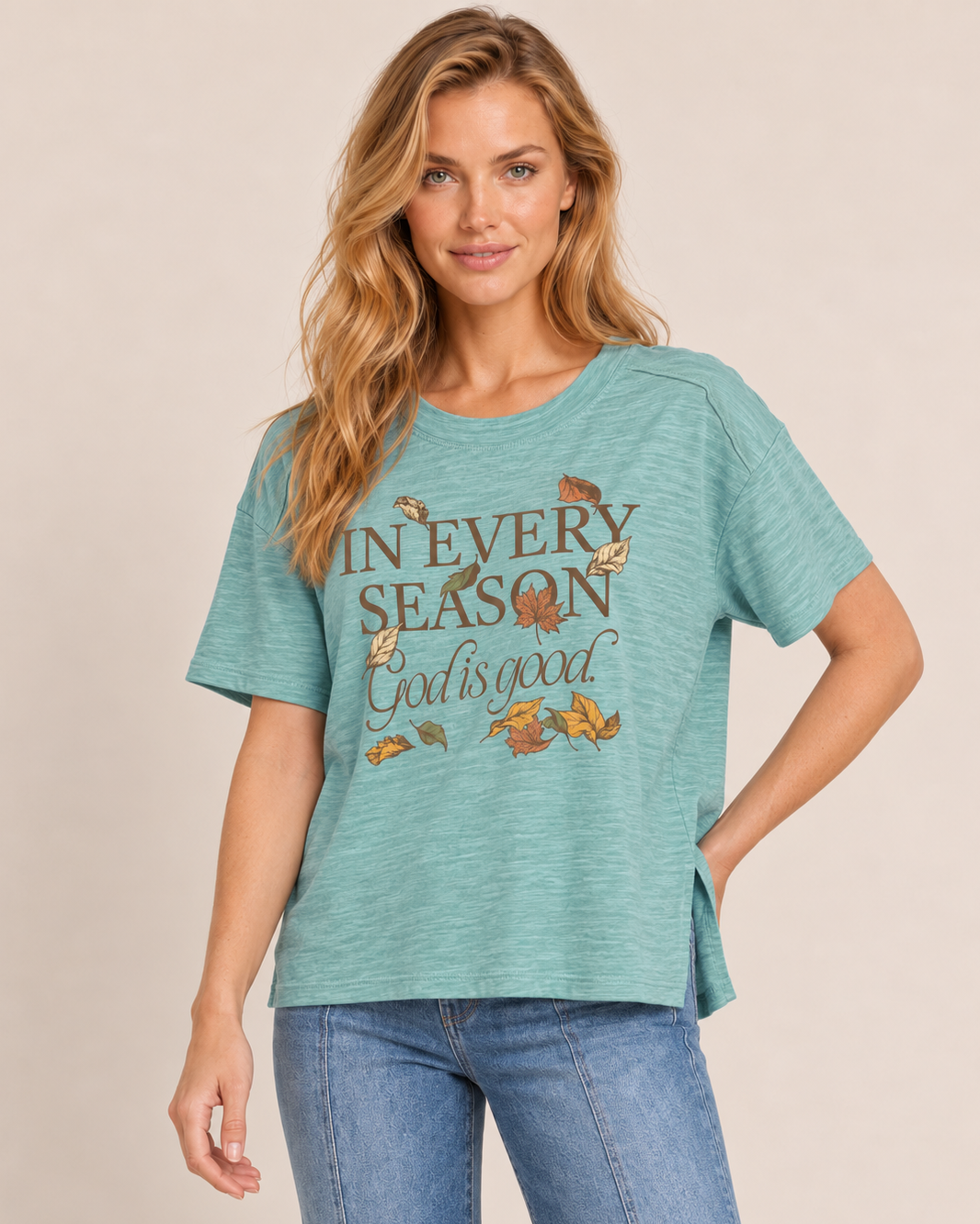 A woman wearing a heathered teal short-sleeve graphic t-shirt with 'IN EVERY SEASON God is good' and autumn leaves printed on the front, paired with blue jeans, against a solid beige background.