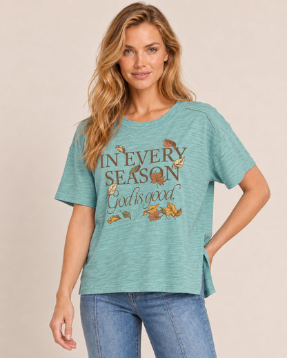 A woman wearing a heathered teal short-sleeve graphic t-shirt with 'IN EVERY SEASON God is good' and autumn leaves printed on the front, paired with blue jeans, against a solid beige background.