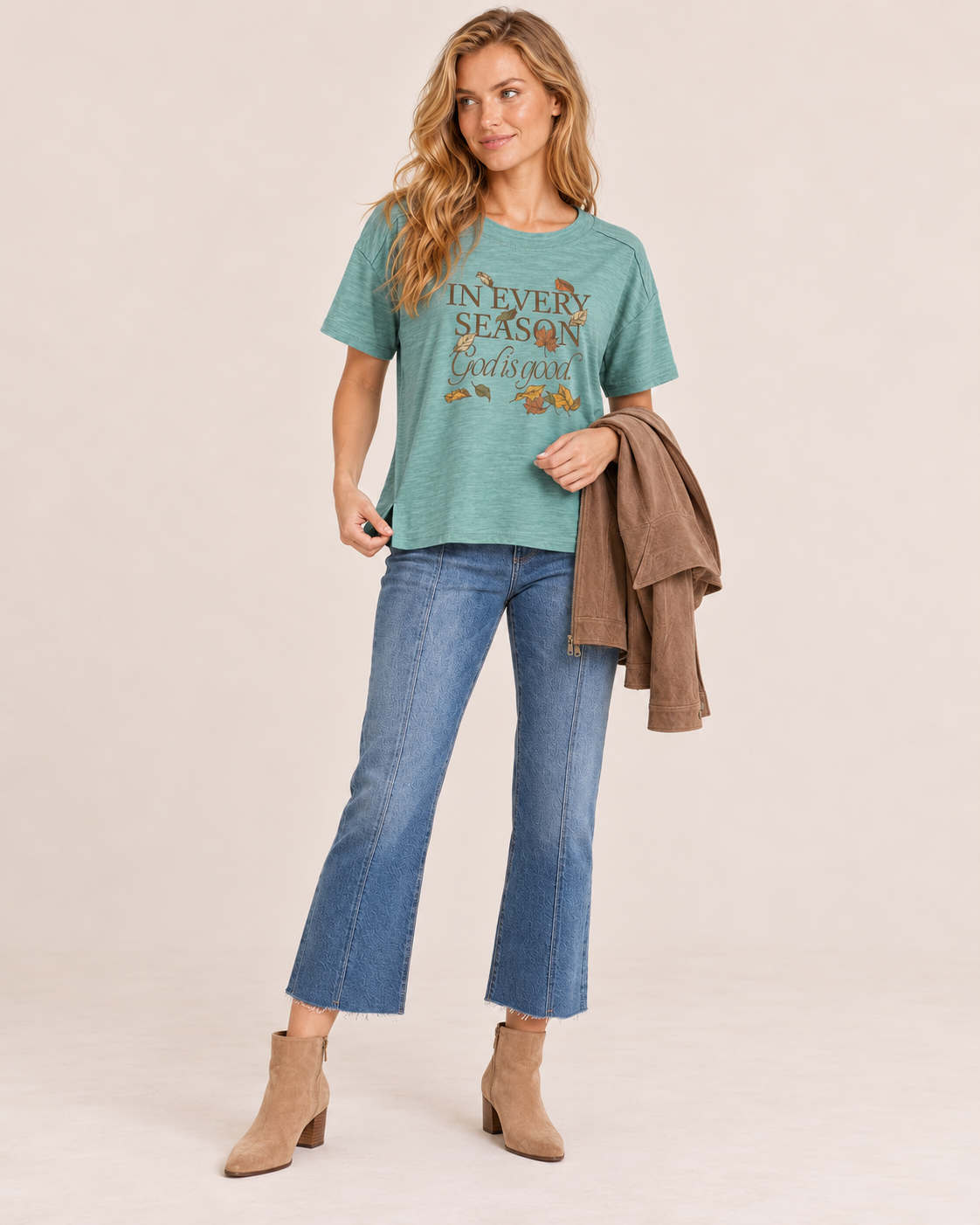 A woman modeling a teal graphic t-shirt with 'IN EVERY SEASON Christmas' and autumn leaf print, paired with blue cropped flare jeans, beige ankle boots, and holding a brown jacket against a solid beige studio background.