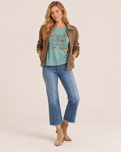 A woman modeling a brown suede jacket over a teal graphic t-shirt, paired with blue cropped jeans and tan ankle boots, against a solid light beige background.