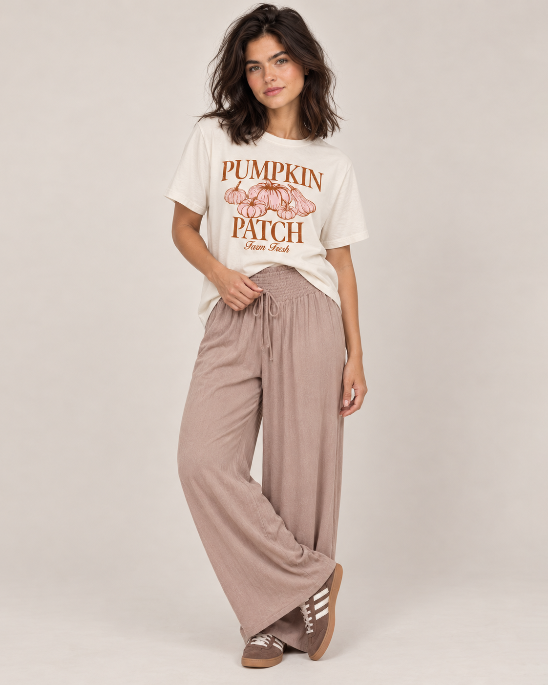 A woman modeling a cream graphic t-shirt with a brown 'Pumpkin Patch' design and matching wide-leg taupe pants, standing against a neutral light beige background.