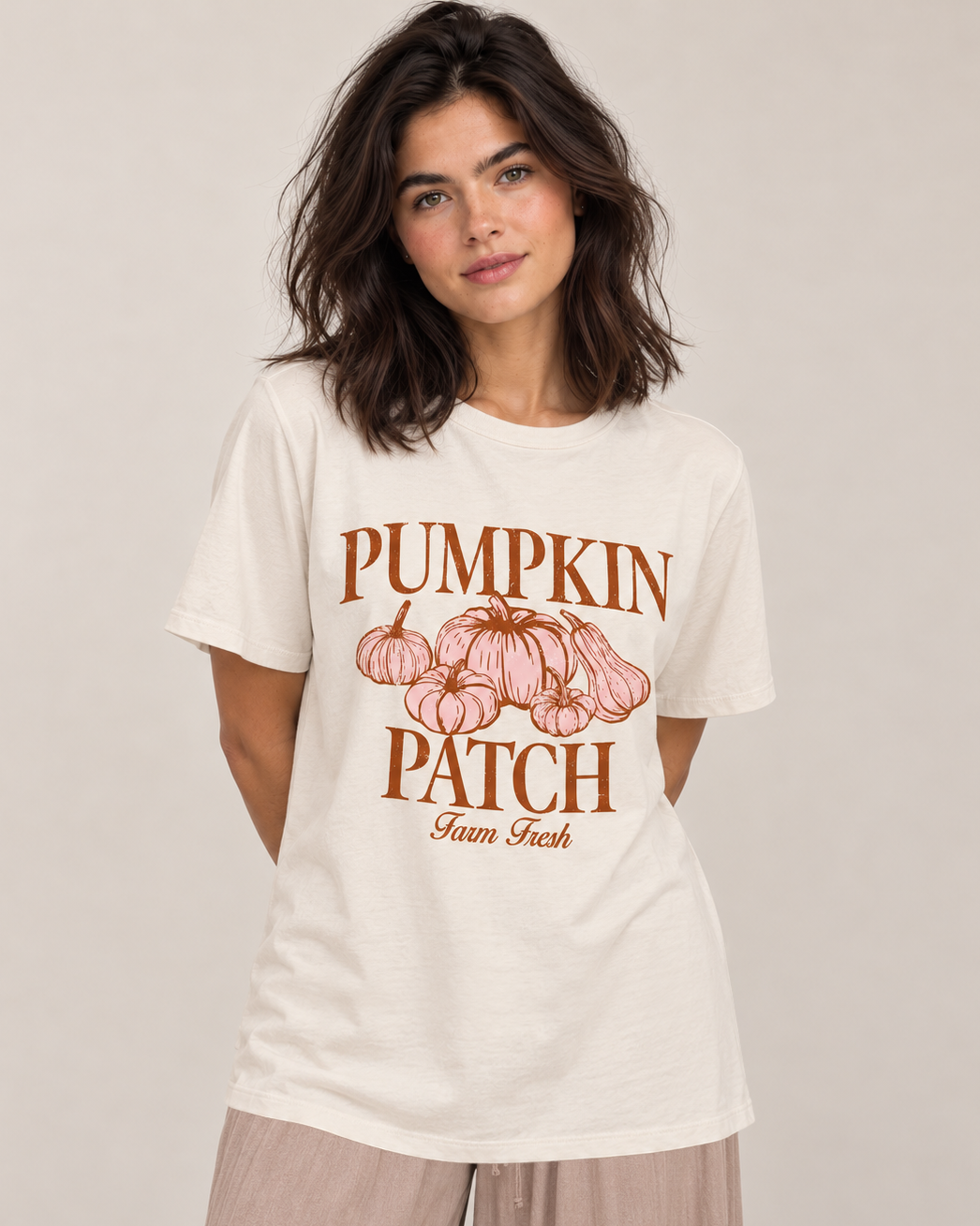A woman wearing a cream-colored graphic t-shirt with a brown and pink 'Pumpkin Patch Farm Fresh' print, paired with light beige pants, against a neutral beige background.