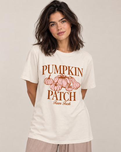 A woman wearing a cream-colored graphic t-shirt with a brown and pink 'Pumpkin Patch Farm Fresh' print, paired with light beige pants, against a neutral beige background.