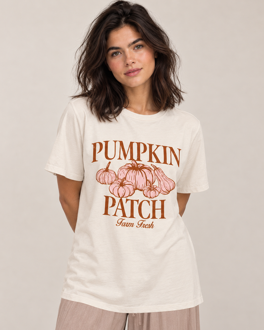 A woman wearing a cream-colored graphic t-shirt with a brown and pink 'Pumpkin Patch Farm Fresh' print, paired with light beige pants, against a neutral beige background.