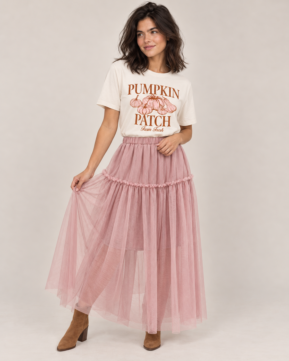 A woman modeling a long, tiered dusty pink tulle maxi skirt paired with a white graphic t-shirt and brown ankle boots, set against a plain light gray studio background.