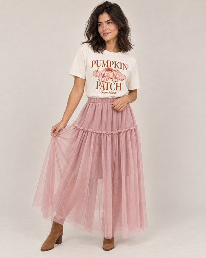 A woman modeling a long, tiered dusty pink tulle maxi skirt paired with a white graphic t-shirt and brown ankle boots, set against a plain light gray studio background.