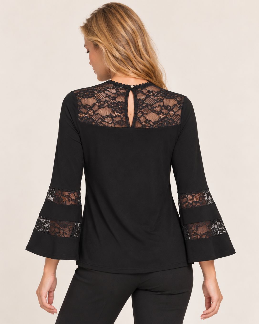 Back view of a woman wearing a black long-sleeve tunic with floral lace sleeve details and black pants on a beige background.