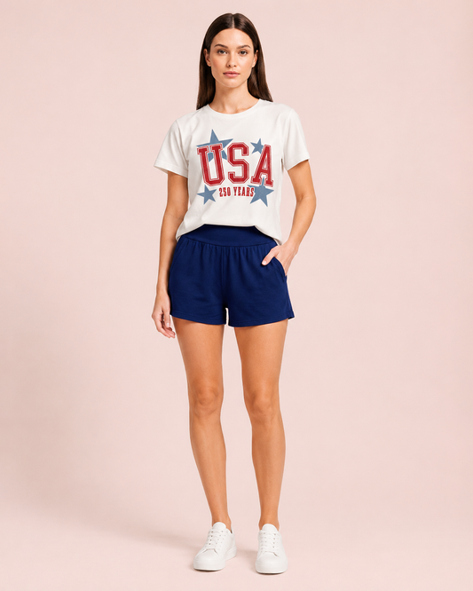 Side view of a woman wearing a white USA graphic tee and navy lounge shorts on a beige background