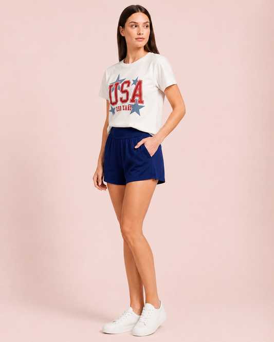 Side view of a woman wearing a white USA graphic tee and navy lounge shorts on a beige background