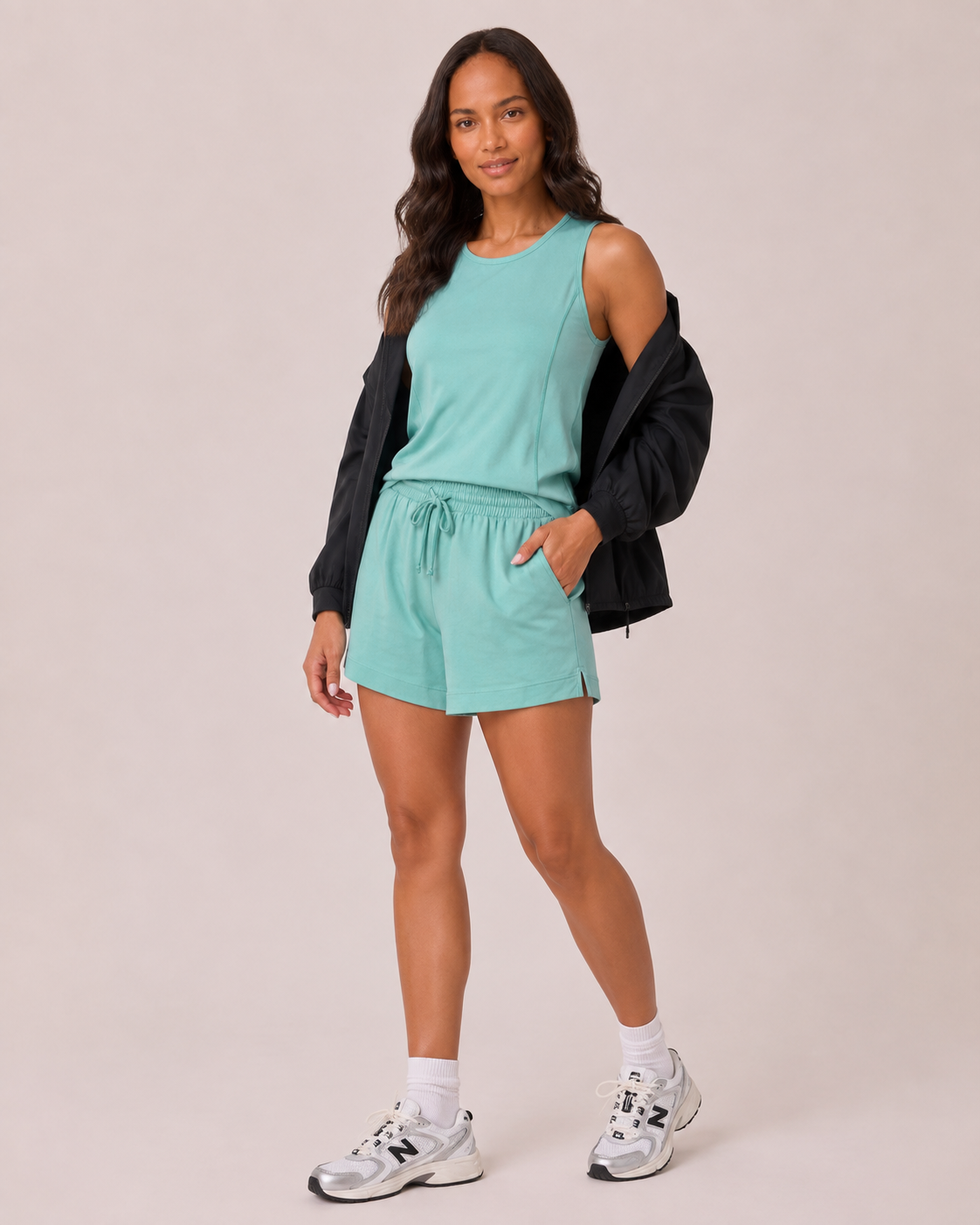 Full-body view of a woman wearing a capri blue tank top with black leggings and white sneakers on a beige background